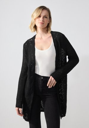 Zwarte, opengehaakte cardigan met lange mouwen en een relaxte pasvorm, gedragen over een witte top, gecombineerd met donkere jeans. Gelaagd patroon zichtbaar.