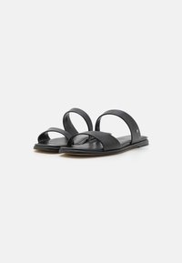 Sandales en cuir noir avec deux larges brides sur le dessus du pied et une semelle plate légèrement courbée. Comprend un petit logo en accent sur le côté.