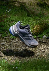 Chaussures d'extérieur grises avec des accents noirs, tige texturée et semelle robuste. Présente un col rembourré et un design à trois bandes. Détail bleu au niveau du talon.