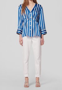 Modrá a bílá pruhovaná blůza s dlouhými rukávy, zapínáním na knoflíky vpředu a výstřihem do V, spárovaná s bílými kalhotami a sandály na průhledném podpatku.