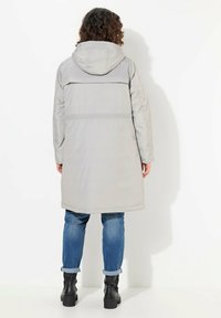 Cappotto lungo grigio chiaro con cappuccio, dotato di una texture liscia, taglio dritto e una cucitura orizzontale vicino alle spalle. Indossato sopra jeans arrotolati e stivali neri.