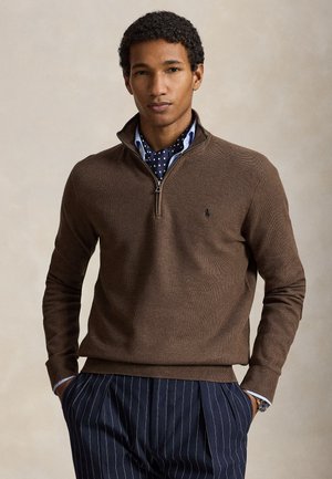 Homme portant un pull marron à fermeture éclair partielle sur une écharpe bleue à pois et un pantalon rayé bleu marine, posant les mains dans les poches devant un fond uni.