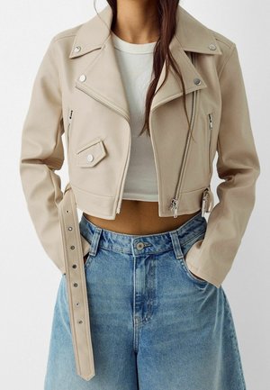 Frau trägt eine beige gekürzte Bikerjacke über einem weißen gerippten Croptop und hellblauen hoch taillierten Jeans mit Händen in den Taschen.