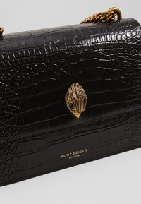 Černá kožená kabelka se strukturou krokodýlí kůže, zlatá kovová spona ve tvaru mušle a řetízkový popruh. Na přední straně je reliéfní logo značky.