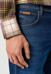 Camisa de cuadros marrón y crema con puño blanco, combinada con unos jeans de mezclilla azul oscuro que tienen una etiqueta de cuero marrón. Mano descansando sobre los jeans.