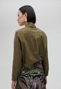 Olive green button-up shirt with long sleeves, featuring black side stripes. The back has a pleat detail for a relaxed fit.
