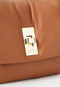 Torba ze skóry w kolorze brązowym z błyszczącą złotą prostokątną klamrą z grawerem "Tommy Hilfiger" na przednim klapie.