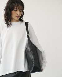 Giovane donna che indossa una camicia bianca a maniche lunghe e una gonna di pelle nera, mentre porta una grande borsa a tracolla nera su uno sfondo semplice.