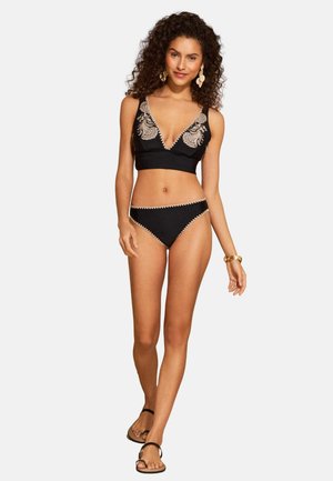 Donna con capelli ricci che indossa un bikini nero con ricamo beige, orecchini d'oro, braccialetto e sandali neri, che cammina con sicurezza.