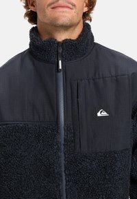 Giacca navy con una sezione inferiore in pile testurizzato, sezione superiore liscia e chiusura con zip. Presenta una tasca sul petto e un accento con il logo.