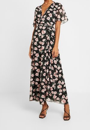 Femme portant une robe portefeuille noire à fleurs roses, manches courtes volantées et sandales à talons noirs à brides.