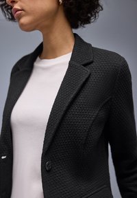 Femme portant un blazer noir texturé sur une chemise blanche unie, montrée des épaules au milieu du torse sur un fond gris.