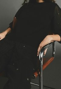 Henkilö istuu ruskeassa nahkatuolissa, jonka käsinojat ovat kromiset, päällään musta kohokuvioitu neulepaita ja mustat housut, joissa on napitusyksityiskohtia.