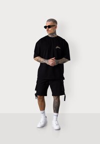 Schwarzes, übergroßes T-Shirt mit einem kleinen goldenen Logo, kombiniert mit schwarzen Cargo-Shorts und weißen Sneakers, die gemusterte Bein-Tattoos aufweisen.