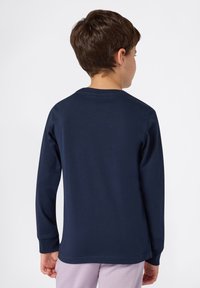 Ein Junge mit kurzen braunen Haaren trägt ein dunkelblaues Langarmshirt und eine helllila Hose und steht mit dem Rücken zu einem weißen Hintergrund.