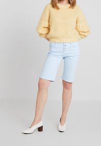 Suéter de punto amarillo claro con mangas farol, combinado con pantalones cortos de mezclilla azul claro y zapatos de tacón blanco.
