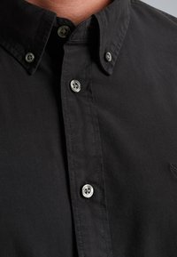 Camicia nera con bottoni realizzata in cotone. Caratterizzata da un collo classico e chiusure con bottoni in metallo argentato. Texture liscia con una leggera lucentezza.