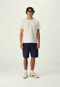 Men's cotton t-shirt in light beige with a chest pocket, paired with navy shorts and cream sneakers featuring red stripes. Sunglasses included.