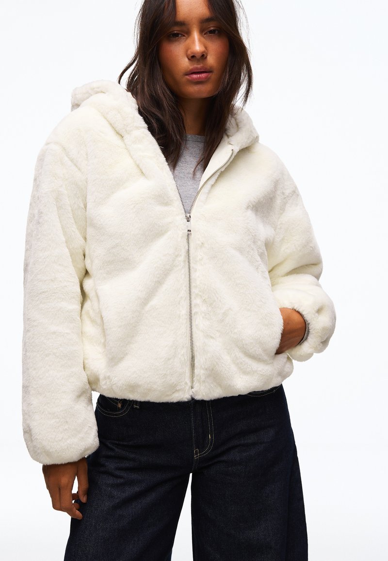 Femme portant une veste blanche duveteuse zippée avec capuche et pantalon foncé, debout avec une main dans la poche de la veste.