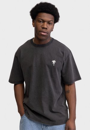 Ung mand med kort afro, iført en vasket sort t-shirt med et lille hvidt palmetræsemblem, lyseblå jeans og en guldkæde.