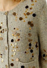 Beige gebreide cardigan met ronde sequins in goud en zwart, met een gestructureerd oppervlak, knopen aan de voorkant en een kleine zak.