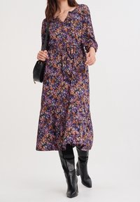 Pletené květinové maxi šaty v purpurové barvě s oranžovými a žlutými akcenty, s výstřihem do V, dlouhými rukávy a páskem v pase, doplněné černými kozačkami do kolen.