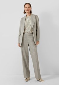 Kobieta w jasnoszarym garniturze z przeskalowanym żakietem i szerokimi spodniami, zestawionym z srebrną, cekinową bluzką i metalicznymi, złotymi butami.