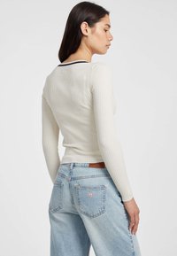 Giovane donna con lunghi capelli scuri che indossa una maglietta bianca a coste con maniche lunghe e jeans a vita alta di colore blu chiaro, girata di lato.
