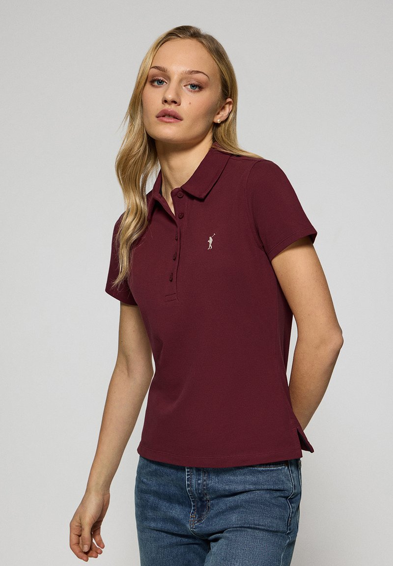 Burgundy pikétröja med krage, fem knappar och en liten broderad logotyp på vänster bröst. Korta ärmar och rak nederkant.