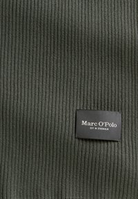 Tissu côtelé vert foncé avec une texture douce. Comprend une étiquette noire avec un texte blanc : « Marc O'Polo EST. IN STOCKHOLM. »