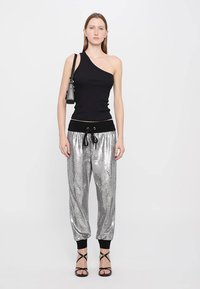 Zwarte top met één schouder, gecombineerd met glanzende zilveren joggers met een trekkoord in de taille en zwarte boorden, aangevuld met zwart, geschoeide hakken.