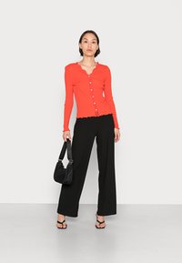 Top de manga longa vermelho com riscas, decote com cascata e detalhes em botão, combinado com calças largas pretas e uma mala preta. Modelo usa sandálias com tiras.