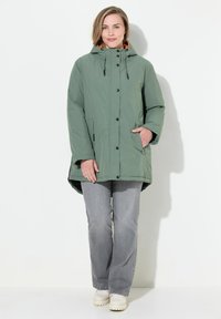 Grön huoodjacka med synliga knappar, en avslappnad passform, två sidofickor och kontrasterande orange foder. Kombinationen med grå jeans och krämfärgade skor.