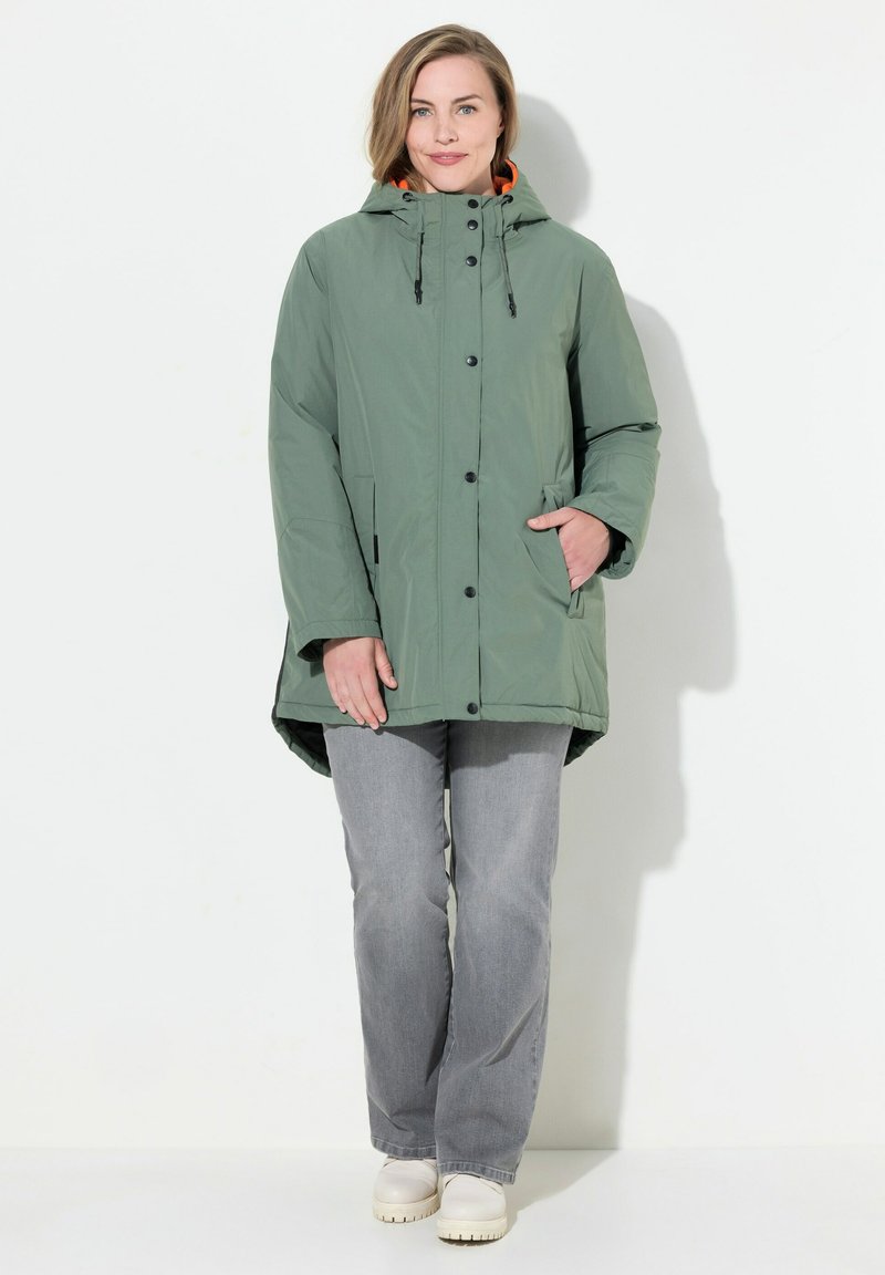 Grön huoodjacka med synliga knappar, en avslappnad passform, två sidofickor och kontrasterande orange foder. Kombinationen med grå jeans och krämfärgade skor.