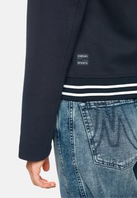 Navy sweatshirt with a fitted cuff, white striped hem, and a branded label. Worn with faded blue denim jeans, showcasing a gradient texture.