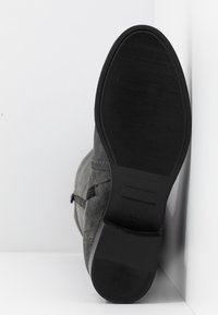 Semelle d'une botte en daim gris avec une semelle en caoutchouc noire, positionnée verticalement contre un coin de mur et de sol blancs.