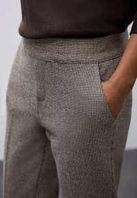 Gros plan sur une personne portant un pantalon à motif pied-de-poule marron, avec une main dans la poche latérale et un pull marron foncé rentré à l'intérieur.