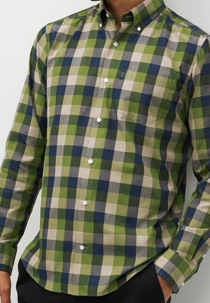 Homme portant une chemise à manches longues boutonnée avec un motif à carreaux vert, marine et beige et une poche poitrine.