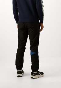 Pantaloni de trening negri cu logo aligator albastru, dungi laterale, manșete elastice și croială tapered. Purtați cu pantofi sport negri și albi.