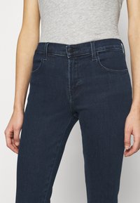 Jeans de mezclilla azul oscuro con corte ajustado, que presentan un cierre de botón, cinco bolsillos y una textura suave. El diseño incluye detalles de costura sutiles.