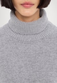 Pull à col roulé gris en maille côtelée épaisse et texturée. Gros plan mettant en valeur la douceur et les détails des coutures.