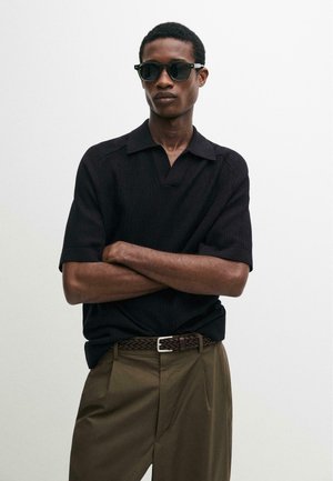 Uomo con camicia nera a maniche corte con colletto, cintura intrecciata marrone, pantaloni oliva e occhiali da sole neri, in piedi con le braccia incrociate davanti a uno sfondo bianco.