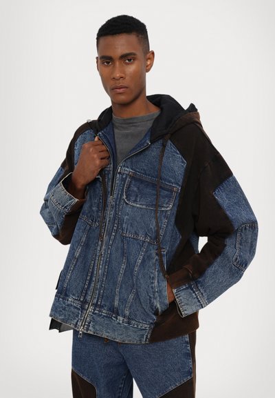 Denim jacket with a mix of dark brown and blue shades, featuring a zip closure, front pockets, and a hood. Texture is rugged and casual.