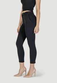 Pantaloni neri aderenti con cintura e passanti per la cintura, dotati di tasche laterali, taglio slim e texture liscia, abbinati a tacchi alti color nude.