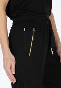 Zwarte joggingbroek met elastische tailleband, voorzien van een gouden ritszak en een klein Zweeds vlagaccent. Gladde stoftextuur.