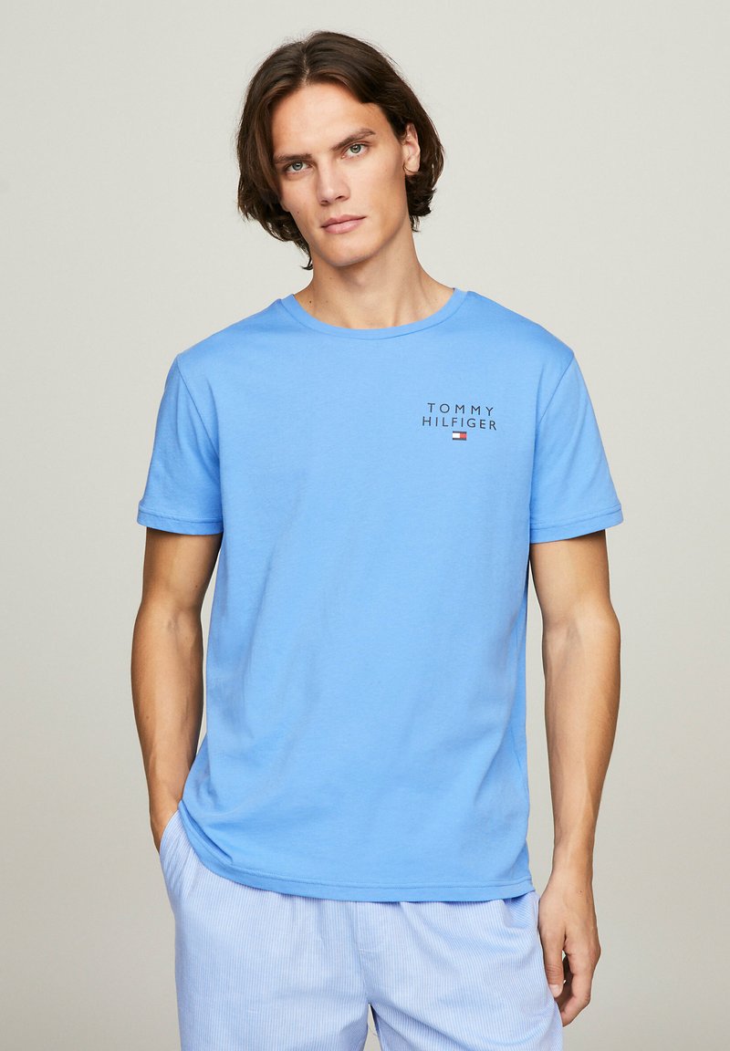 T-shirt di cotone azzurro chiaro con maniche corte. Presenta il testo "TOMMY HILFIGER" e un piccolo logo nell'angolo in alto a sinistra.