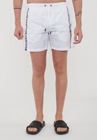 Pantaloni da bagno bianchi con dettagli neri sui lati e cordino regolabile. Indossati con ciabatte nere, dotate di suola piatta e superficie testurizzata.