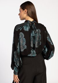 Zwarte blouse met teal paisleypatroon, ballonnenmouwen en een strikdetail aan de achterkant van de hals. De stof lijkt lichtgewicht en iets doorschijnend.