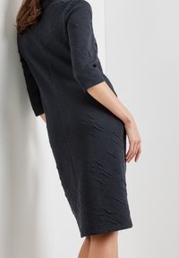 Abito aderente blu navy con tessuto testurizzato; presenta maniche a tre quarti, lunghezza fino al ginocchio, motivo sottile e cuciture che creano una silhouette su misura.