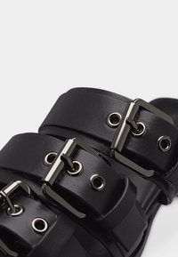 Κοντινό πλάνο μαύρων δερμάτινων σανδαλιών με δύο φαρδιές λουρίδες που φέρουν ασημένιες ορθογώνιες μεταλλικές πόρπες και μεταλλικά οπές.